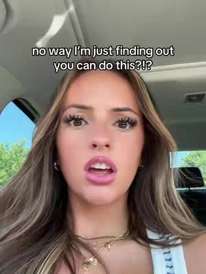 A selfie shot of a young woman with long brown hair, looking at the camera with a surprised expression. She has long eyelashes and is wearing a tank top and a gold necklace. The background shows a vehicle interior, and an overlay of text reads, "no way I'm just finding out you can do this?!"
