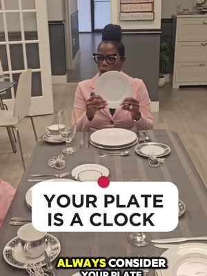 A woman wearing glasses and a pink blazer holds up a white plate in front of a table set for dining. The table is covered with a gray tablecloth, and there are settings for two people, including silverware, glasses, and small plates. The background includes a wall with decorative panels and a door. Text overlays read "YOUR PLATE IS A CLOCK" and "ALWAYS CONSIDER YOUR PLATE".
