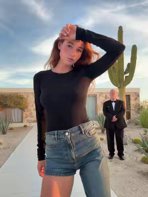 A woman with auburn hair stands in front of a desert backdrop, a hand raised to her forehead. She wears a black long-sleeved shirt and blue jeans. In the background, a middle-aged man in a tuxedo stands on a walkway. The scene is framed by a large cactus and a modern building.