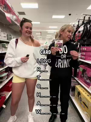 Two young women are standing in what appears to be a store, with two rows of shelves behind them. The woman on the left wears a white sweatshirt and yellow shorts; she has sunglasses on top of her head. The woman on the right is wearing a black hoodie. On the image, there is the text of a game is visible.