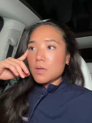 A woman is seated inside a car, likely a Tesla, with the camera framing her face close up. She has long, dark hair and is wearing a dark colored jacket. She has a thoughtful expression, resting her index finger against her temple as if pondering. The lighting seems natural, possibly daylight, with the car's interior visible around her in soft focus.