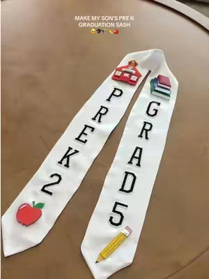 A handmade graduation sash featuring the words 'PREK' and 'GRAD', along with school-themed embellishments. The sash is white, with black text and colorful illustrations, set against a brown background.