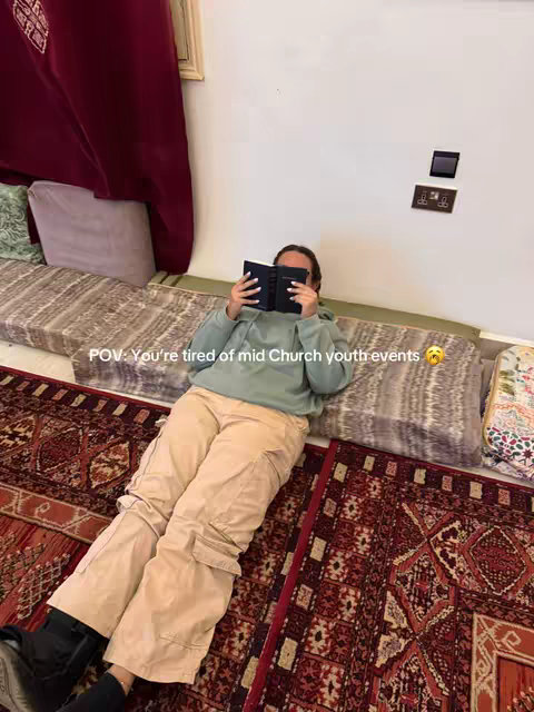 A person is lying down, covering the face with a book. The person is wearing a light green hoodie, a pair of tan cargo pants, and black boots. It's set in front of a white wall, with a burgundy curtain and a multi-colored rug in the foreground.