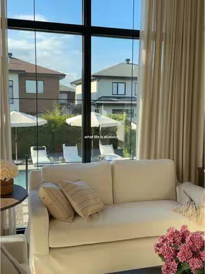 A well-lit interior scene featuring a cream-colored sofa with two decorative pillows. A large window behind the sofa frames the exterior with views of houses, a pool, and umbrellas. The overall composition is clean and modern.
