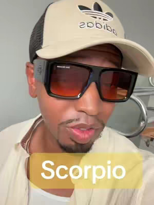 A man wearing a beige Adidas cap and dark sunglasses is the central focus. The background is neutral, with a subtle glimpse of a metallic structure. A text overlay with the word "Scorpio" is placed at the bottom, using a soft yellow backdrop.