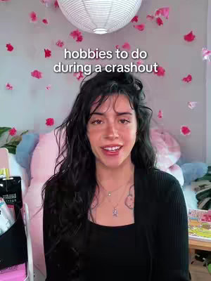 A woman with dark hair and a black top smiles at the camera, with text that reads, “hobbies to do during a crashout.” The background includes a white paper lantern, pink paper decorations against a light-colored wall, and a pillow.