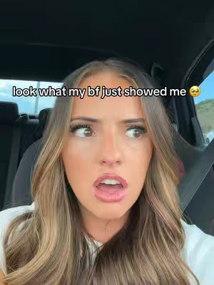 A woman with long, light brown hair is seated in a car, turned with a look of surprise and slight disbelief while facing towards the camera, and there's white text overlaid on the top left. The background shows the inside of the car and an out-of-focus outdoor scene.
