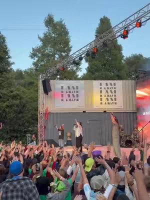 The cover image shows a stage set up outdoors, likely at a camp or similar event. The stage has a simple backdrop with text, and overhead lighting rigs. Performances are taking place on the stage. A large crowd with their arms raised is in front of the stage. The scene suggests a live performance at night, with stage lighting creating a vibrant atmosphere.