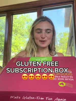A woman is in front of a window showing a green scene. The woman is holding a magenta subscription box that has text overlaying it. The text says: GLUTEN FREE SUBSCRIPTION BOX with six hearts. The box is facing the camera and the woman is looking at the camera.