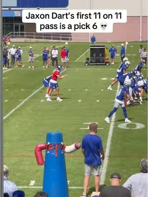 A live-action shot on a green football field during practice. A player dressed in red throws the ball. In the middle of the field, there's a blue inflatable practice dummy next to the person filming the video. Different players wearing blue uniforms stand in the background. The sky is blue, and the lighting is bright.