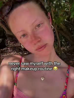 A selfie-style shot of a young woman with fair skin, sunglasses on her head, wearing a pink floral top, and a necklace. She's outdoors, with a natural backdrop of trees, leaves, and sunlight. Text overlay, in white font, reads, 'never saw myself with the right makeup routine'.