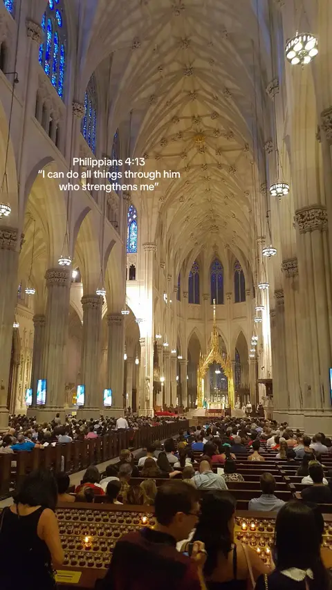 The image is a vertical shot inside a cathedral. The composition features a high, vaulted ceiling with ornate architectural details. Sunlight streams through the stained-glass windows, illuminating the interior. Below, rows of people are seated, likely attending a service. The overall impression is one of grandeur, spirituality, and community.