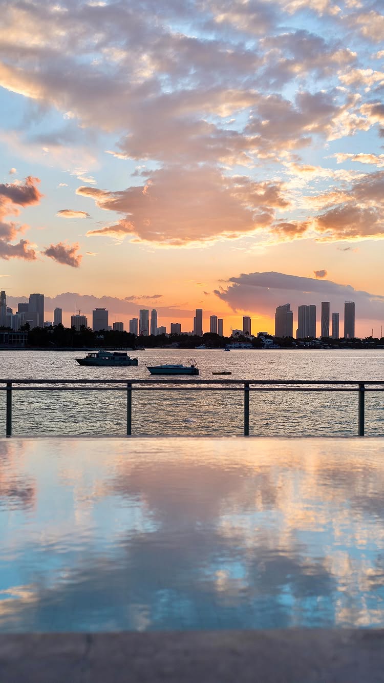 A serene image capturing a sunset over a cityscape and water. The sky is filled with colorful clouds ranging from blues to oranges and pinks, reflecting on the water's surface. Silhouetted buildings form a skyline along the horizon. Two boats are visible on the water. The foreground features a reflective surface, likely a pool, creating a mirror-like effect to the view.
