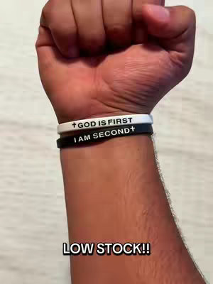 A close-up shot showcases a person's forearm and clenched fist. Three wristbands are worn, with the top one white, the middle one black, and the bottom one obscured. Text overlays on the bands read, 'GOD IS FIRST' and 'I AM SECOND'. The bottom of the frame features an additional text overlay that reads, 'LOW STOCK!!'