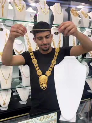 A man poses with a large gold necklace in a jewelry store displaying gold necklaces and jewelry in the background.
