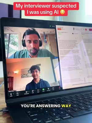 A laptop screen displays a video call with two participants; the person at the bottom is younger, and the person at the top is an older gentleman wearing headphones; above the gentleman, a red banner states 'My interviewer suspected I was using AI'. Below on the laptop screen in large yellow letters says 'YOU'RE ANSWERING WAY'. The background of the image is comprised of walls and various indoor objects.