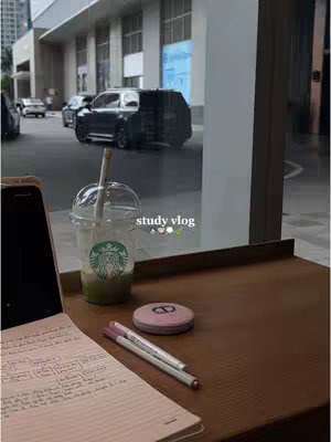 The image captures a study setup in front of a large window overlooking a street scene. On a wooden table, there's a clear plastic Starbucks cup with a green beverage and a straw, alongside a notebook with some writings, two pens and a pink compact. The window reflects the city outside, with buildings and cars.