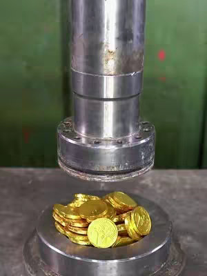 Close up shot of a silver metal object pressing down on a pile of golden coins. The background is a solid green color.