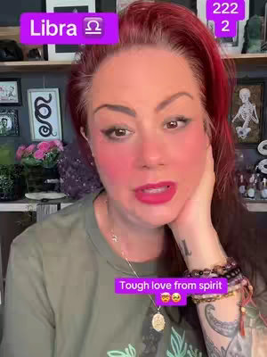 A woman with red highlights in her hair is centered in the frame, looking at the camera. Pink blush is visible on her cheeks and lips. She wears a green top and jewelry, with a tattoo on her arm. The background includes a shelf with various decorations and artworks. Text boxes with purple backgrounds overlay the image, one with the word "Libra" and another stating "Tough love from spirit".