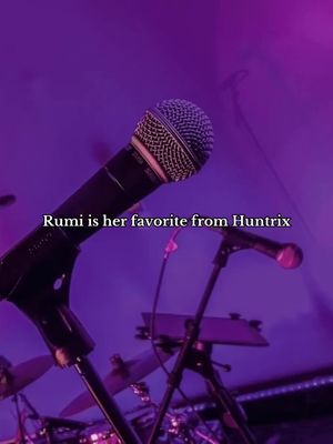 A vertical image featuring a microphone in the foreground, angled to the left, against a purple-lit background. Behind the microphone, there are additional microphone stands and blurry shapes that could be either band members or other stage equipment. The text overlay reads 'Rumi is her favorite from Huntrix'.