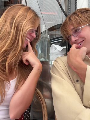 Two young people are seated at a table, likely at a cafe or restaurant, and interacting intimately. A woman with long, light-colored hair and glasses is on the left. She is smiling widely, with her index finger touching her lower teeth. On the right, a man with light brown hair and glasses, wearing a beige jacket, appears to be smiling as well. The background is slightly blurred, suggesting an outdoor setting with architectural details.