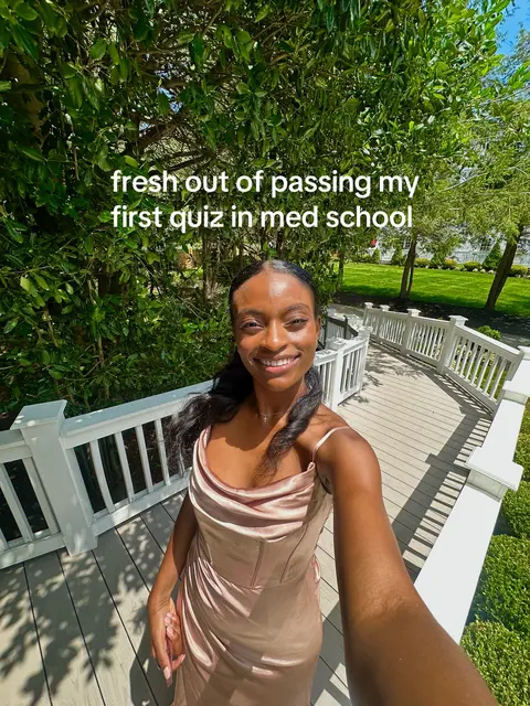 A selfie-style photo shows a young woman smiling towards the camera. She is in a dress that appears to be satin. Behind her, are a series of white railings leading down some stairs from where she seems to be standing. There is an abundant amount of green foliage and trees in the background, suggesting an outdoor setting on a sunny day.