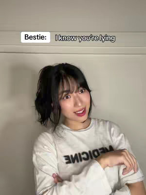A young woman with black hair and bangs is posed at a slight angle, looking towards the right with a slight smile. She's wearing a white t-shirt with text across the front. Above her head, in a white rectangle, is the text 'Bestie: I know you're lying'. The background is a plain white wall.