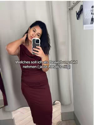 A person is taking a mirror selfie in a dressing room. The person is holding a phone, and also in full mid body shot with a maroon dress. The background consists of basic clothing items and a sign, the person is standing in a stall.