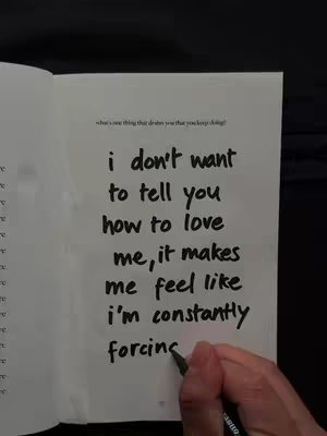 The image shows an open notebook on a dark surface. The notebook has white pages with handwritten text in a bold, black marker. A hand holding a pen appears to be in the process of writing the last word of the last sentence.