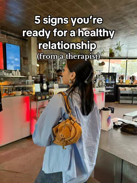 A woman stands in a cafe, seen from behind, looking towards the right side of the frame. She wears a light blue button-down shirt, her dark hair is pulled back with a clip, and she carries a tan purse. The background consists of a cafe interior with counters, shelves, and other customers. The hook text is overlaid at the top.