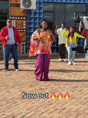 The cover image shows a group of people, apparently in a dance or music video setting, in front of a building with colorful design features. The foreground features a woman in a colorful outfit and purple pants as the central figure. Two men and another woman are in the background, also wearing bright attire and possibly performing, and the words "Now out" accompanied by flame emojis are superimposed.