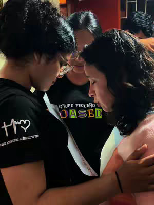 Three women are closely clustered, heads bowed in a gesture of prayer or contemplation, with a sheet of paper in their hands. They are indoors, and the lighting is dim, with the women's faces partially obscured by shadow and hair. The woman in the foreground on the left is wearing a black t-shirt with a heart and a design, while the woman in the center has glasses and a t-shirt with colorful text. The text on the t-shirt appear to belong to a local group