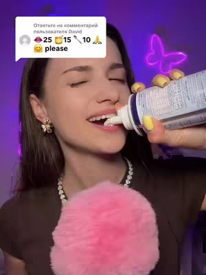 A woman with long brown hair is the central focus, smiling as she dispenses whipped cream from a can directly into her mouth. Her head is tilted back slightly. She has visible makeup and is wearing a dark brown top. A pink fluffball is held near her chest, and a string of pearls is around her neck. Behind her, to the right, a neon butterfly is visible. A comment box floats above her head, showcasing a user's comment.
