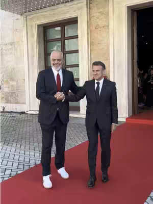 Two men in suits shaking hands on a red carpet in front of a building with large windows.