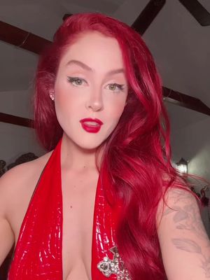 A woman with red hair and red lipstick, wearing a red halter top, looks toward the camera. She has fair skin, with visible makeup including eyeliner, eyeshadow, and blush. Her red hair is styled with volume. The background is blurred, suggesting an indoor setting.