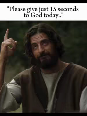 A man with a beard, long hair, and a brown tunic is pointing upwards with his right index finger. The image has a blurred background of trees, suggesting an outdoor setting.