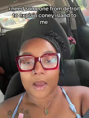 A woman with dark hair in braids and large red-framed glasses is positioned inside a car, facing the camera. Text overlays at the top of the screen. The background shows a bit of the car's interior out of focus.