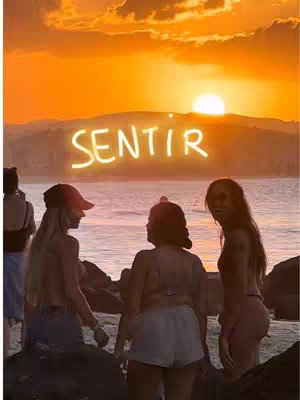 The cover photo features a sunset scene at the beach. The sky is filled with warm orange and yellow hues, and the sun is setting over the water. Silhouetted figures of three women stand on the shore, appearing to be in conversation. Above them, the word "SENTIR" is written in glowing, stylized letters.
