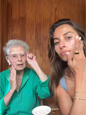 A selfie-style video featuring a young woman and her grandmother. The young woman, on the right, applies skincare product. The grandmother, on the left, is wearing glasses, and a green top, and holding her face with her hands. Both are in front of a wooden background.