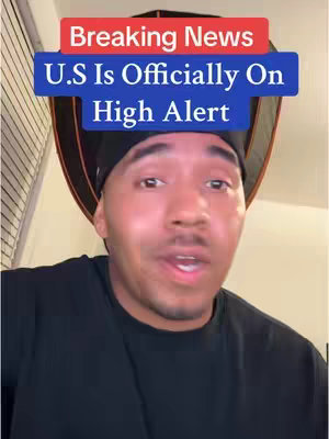 A man with dark hair and a black shirt is centered in the frame, appearing to be speaking. A text overlay above his head reads "Breaking News," with the text "U.S Is Officially On High Alert" beneath. The background is a mix of a black umbrella-shaped element and warm ambient lighting.