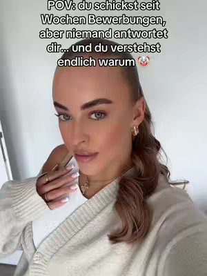 A woman is in a selfie-style video, close up, leaning slightly towards the camera, with text overlaid on top. She has light skin, styled brown hair swept to one side, wearing a cream-colored sweater. The background is simple and light, with a glimpse of a white object on the left side of the frame.