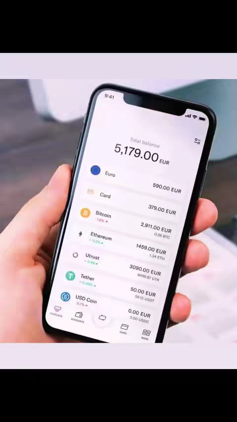 An iPhone displays a financial app interface. The phone is held in a hand, with the screen showing a breakdown of assets including Euro, Bitcoin, and Ethereum. The screen's color scheme is primarily white and blue.