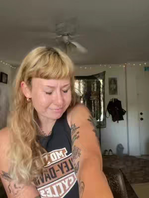 A woman with blonde hair and a slight smile is the central focus, viewed from the chest up. She appears in a well-lit indoor setting, likely a home, with a ceiling fan visible above. She has a few tattoos, is wearing a tank top, with her face showing slight discomfort..