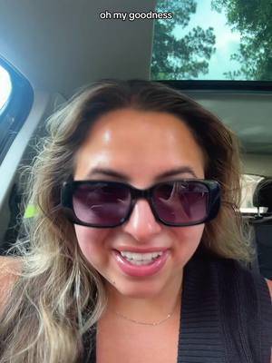 A selfie-style image features a woman seated inside a car, with the camera positioned to capture a close-up of her face. She wears large, rectangular sunglasses that reflect the interior and exterior of the vehicle. Her wavy, light-brown hair frames her face, and she is smiling, with visible teeth, suggesting a friendly or joyful expression. Behind her, a partially visible window shows a blurred view of trees, and details of the car's interior are present. The overall lighting seems natural.