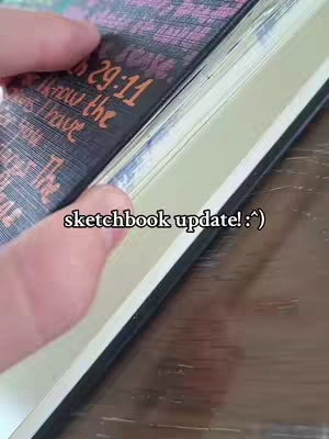 A hand holding open a sketchbook, revealing a page with text and the corner of a black book cover.
