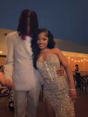 A well-lit photo captures a couple at an outdoor event, likely a formal gathering. The woman, in a sparkling gold gown with a high slit, embraces the man in a light-colored suit. The background shows an evening scene with dim lighting and blurred figures, contributing to a soft focus. The composition highlights the couple, emphasizing their attire and embrace.