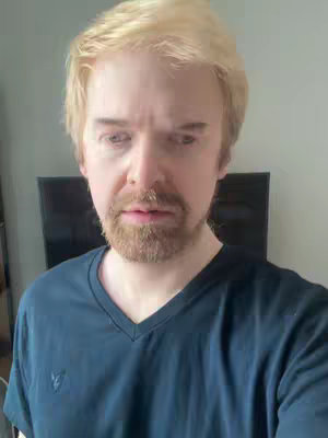 A medium shot shows a man with short, light-colored hair and a neatly trimmed beard. He wears a navy blue V-neck shirt. The background is a light gray wall, and a dark screen, possibly a television, is visible behind him.