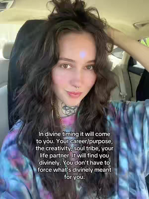 A woman with long wavy dark brown hair sits in a car. Text overlay is in a white box at the bottom of the image. The woman is wearing a blue and purple tie-dye shirt. A purple dot is on her forehead. She has tattoos on her neck and a nose ring, and she is looking directly at the camera with a slight smile.