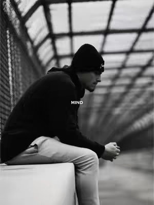 A black and white photo of a man in a black beanie and hoodie, sitting pensively. He is sitting on a white surface with a chain-link fence and metal structure in the background.