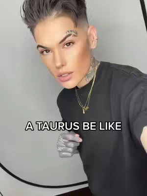 A man with styled dark hair and a face tattoo stands with a neutral expression. He is wearing a black t-shirt and a necklace. Text overlayed on the image reads 'A TAURUS BE LIKE'.